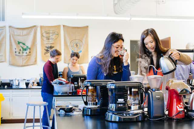 In a lab classroom, a student sips a glass cup of coffee while another pours from a gooseneck kettle and steam rises through the air. The tables have coffee makers and grinders on them. Two students stand over a coffee maker in the background. 