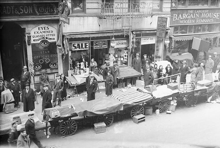 A black and white photo shows a row of pushcarts on a sidewalk, their wares covered up by cloths.