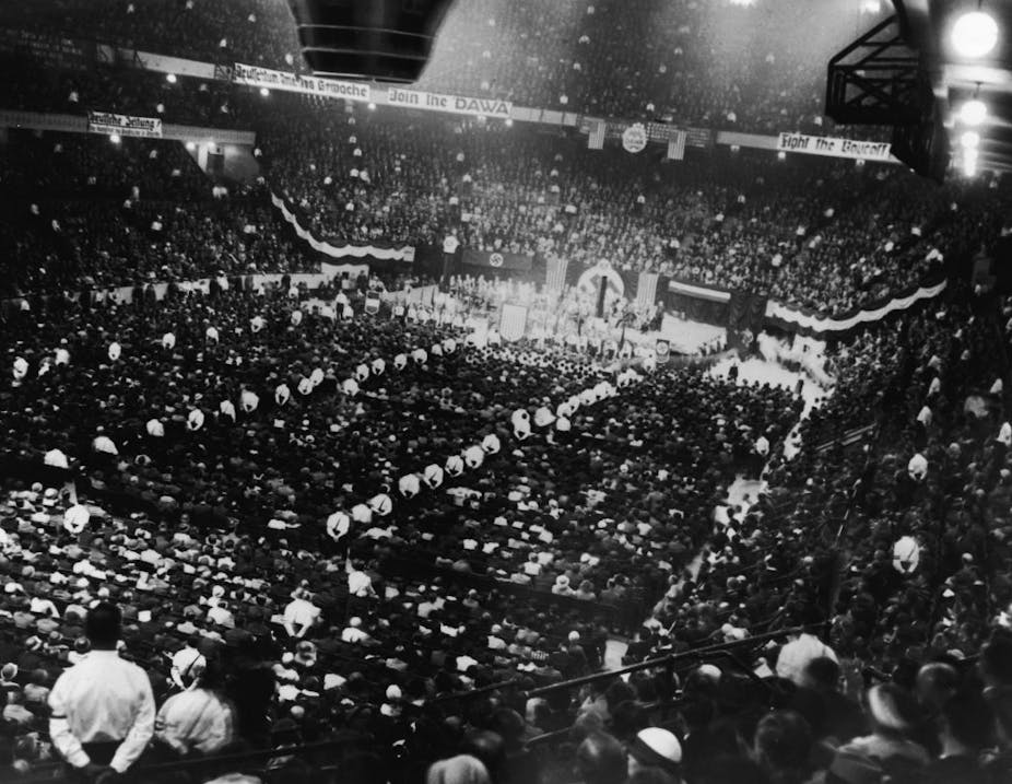 A black and white photo from the back of a huge hall with large crowds of people assembled and looking toward the stage.