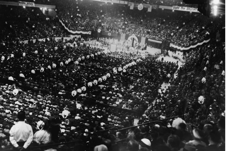 A black and white photo from the back of a huge hall with large crowds of people assembled and looking toward the stage.