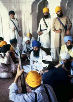 Several men, wearing yellow or blue turbans and flowing white shirts, standing inside a building, while holding guns.