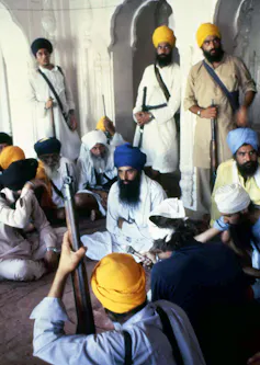 Several men, wearing yellow or blue turbans and flowing white shirts, standing inside a building, while holding guns.