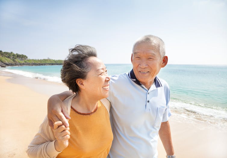 Older Asian couple happily walking along beach, looking at each other