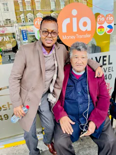 Two men pose for a photo in front of a shop, a younger man standing with his arm around a seated elder man.