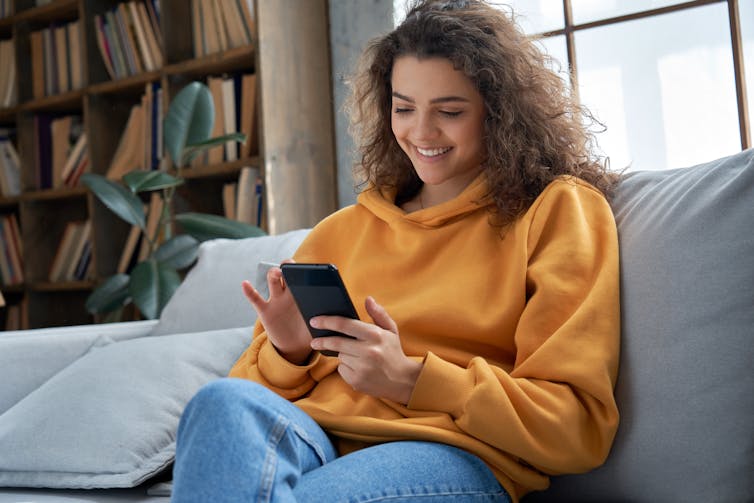 Young woman in a yellow hoodie smiling at her phone.