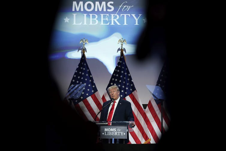 A man delivers remarks with a Moms for Liberty sign behind him.