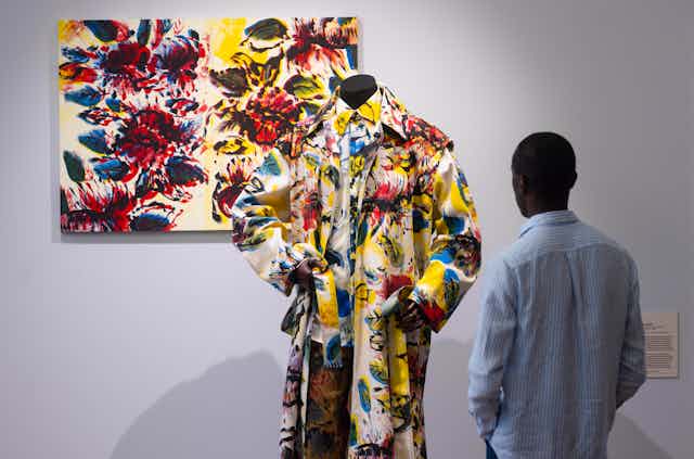 A paint splattered suit in front of a matching canvas, with a young black man looking on