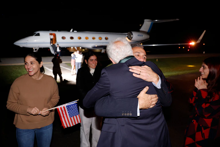 Two men hug one another at an airport while three smiling women look on.