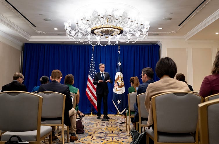 A man, dressed in a blue suit and tie, speaking to a group of people.