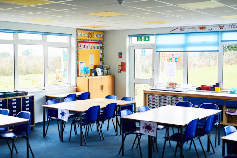 A classroom with tables and chairs . There are two banks of windows and a wood cabinet in the corner with a globe sitting on top.