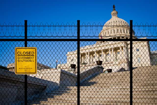 A padlocked gate topped with barbed wire is seen in front of the U.S. Capitol building.  A sign on the fence reads "CLOSED GOVERNMENT SHUTDOWN"