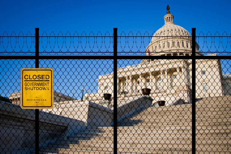 A padlocked gate topped with barbed wire is seen in front of the U.S. Capitol building. A sign on the fence reads "CLOSED GOVERNMENT SHUTDOWN"