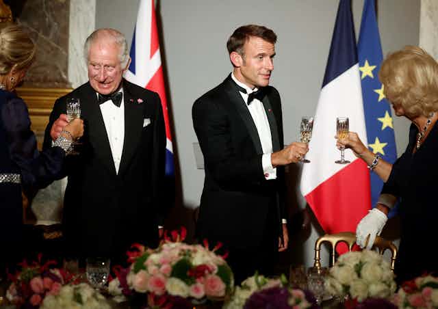 King Charles and Emmanuel Macron clinking glasses with each others wives at a black tie banquet. 