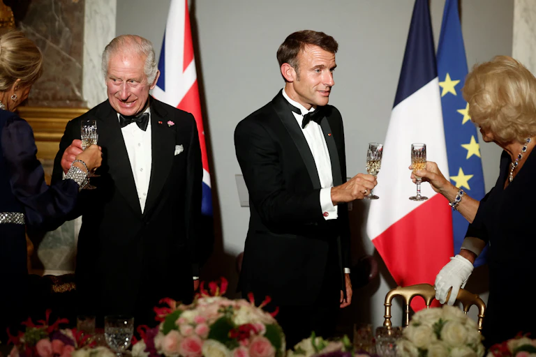 King Charles and Emmanuel Macron clinking glasses with each others wives at a black tie banquet.