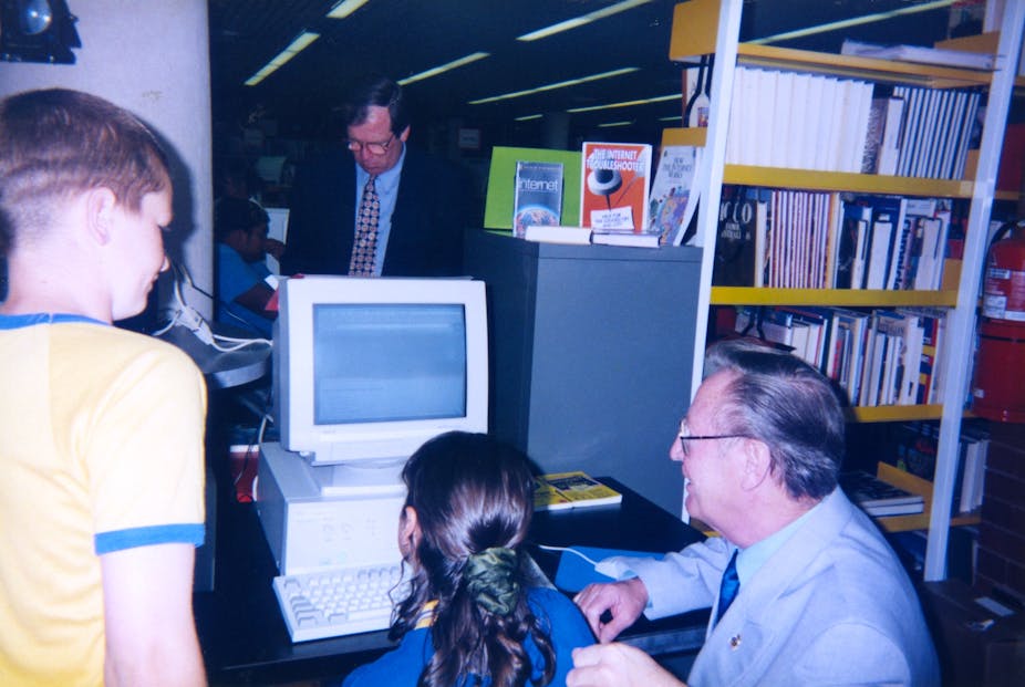 An old man and two children using an old beige computer in a library.