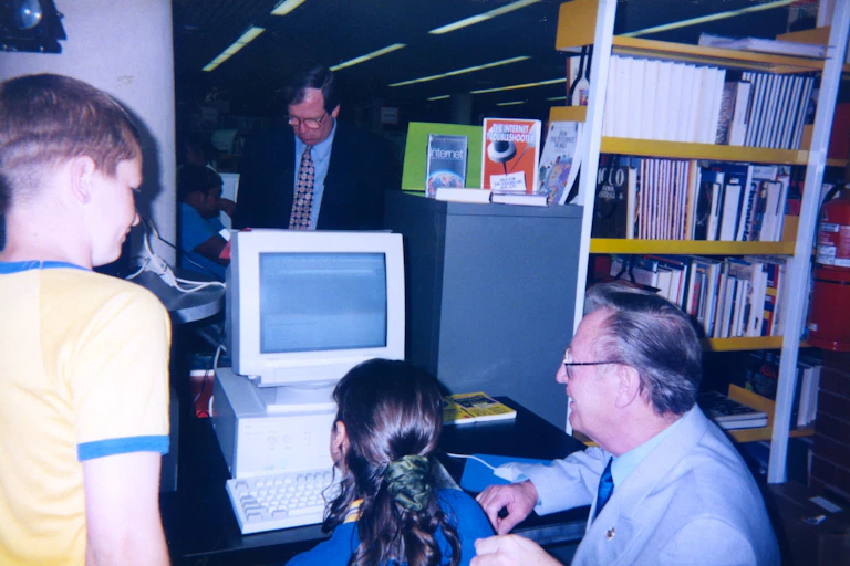 An old man and two children using an old beige computer in a library.
