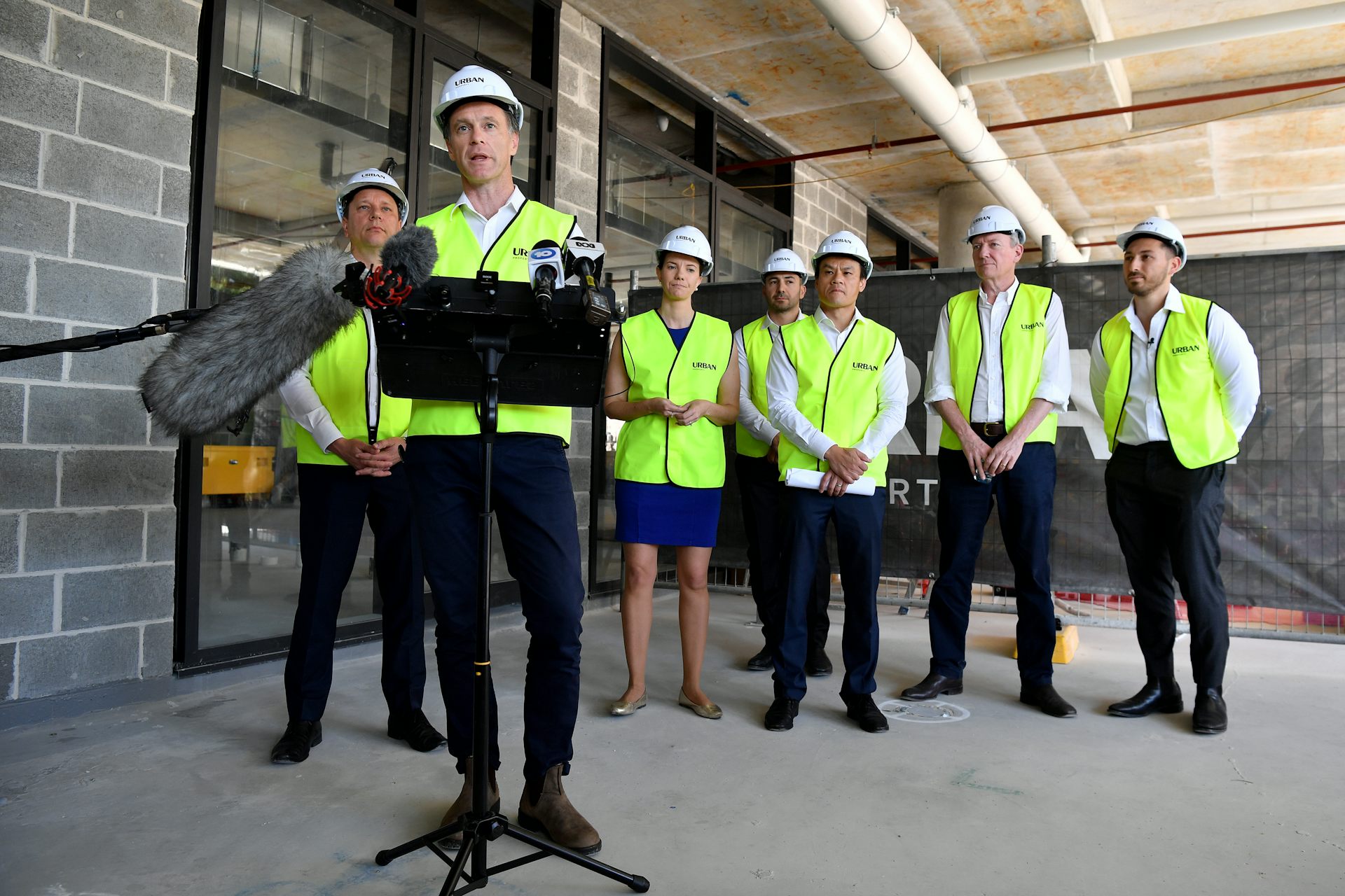 Line of people stand behind a man at a lectern – all wearing hard hats and high-vis – at an apartment development site