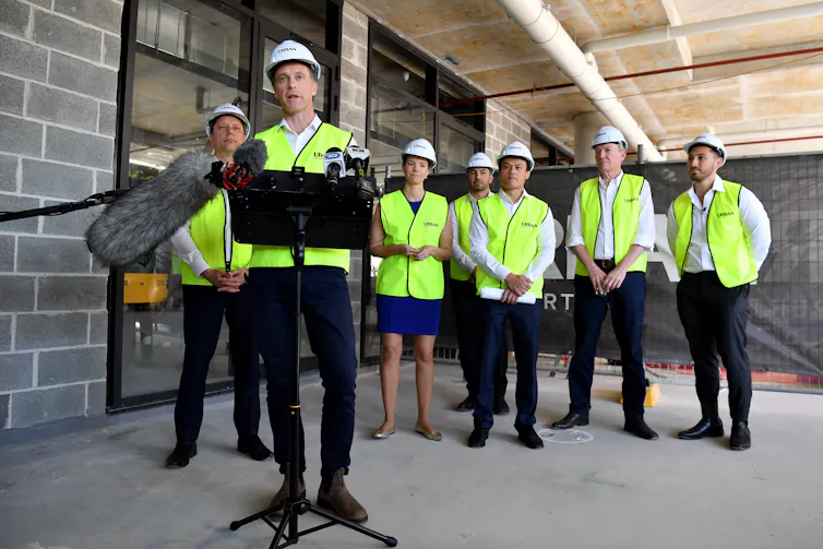 Line of people stand behind a man at a lectern – all wearing hard hats and high-vis – at an apartment development site