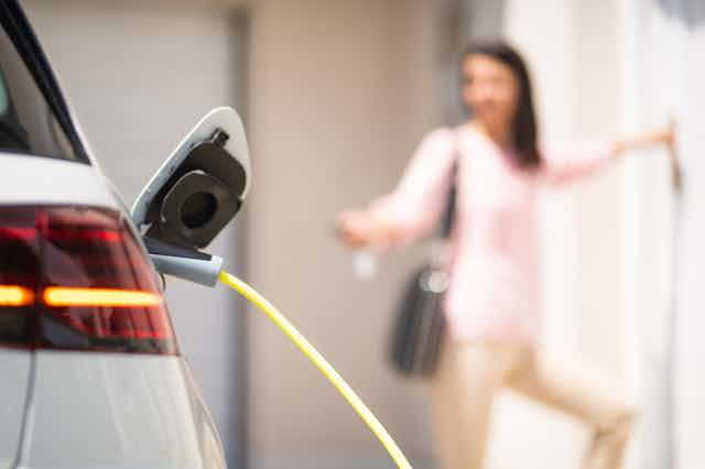 A person out of focus locks the door of their charging electric car.