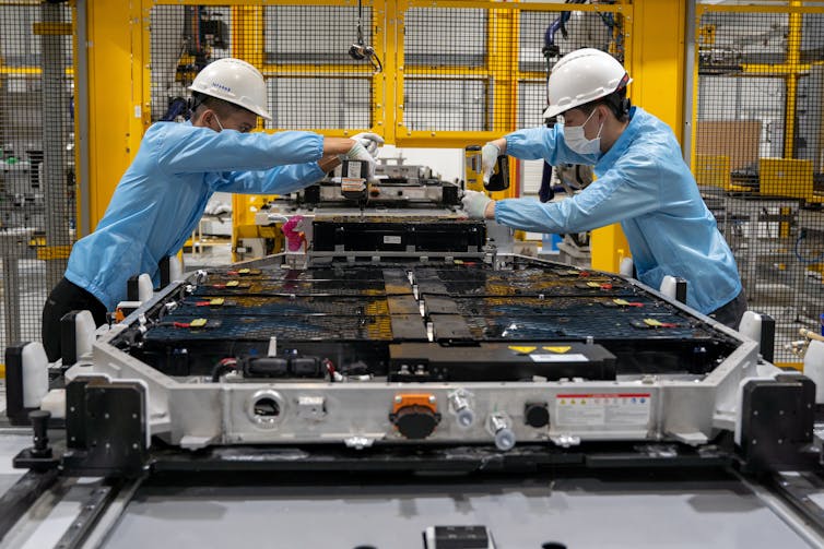 Two workers lean over an EV battery pack on a factory assembly line.