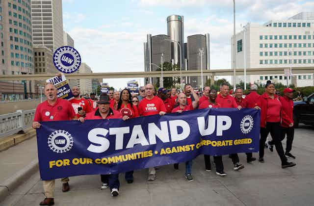 Men and women dressed in red with UAW logos march while carrying a banner that says, 'Stand up for our communities against corporate greed.'