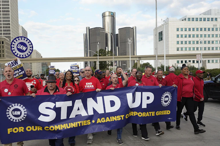 Men and women dressed in red with UAW logos march while carrying a banner that says, 'Stand up for our communities against corporate greed.'