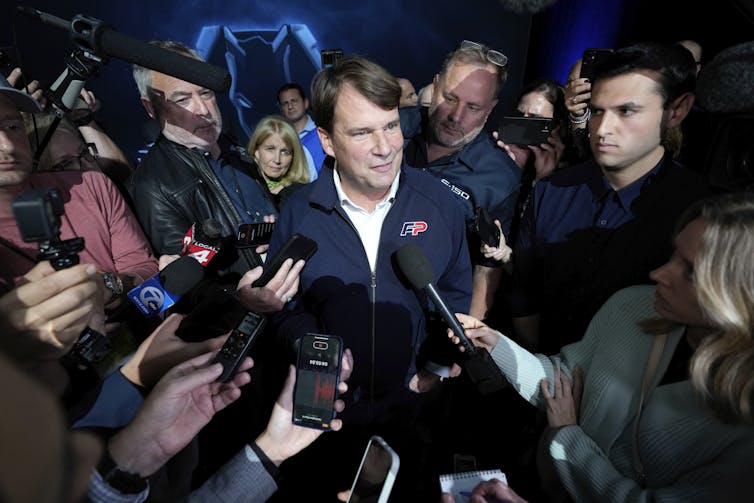 Man in a blue jacket and white t-shirt surrounded by journalists holding microphones and recording devices