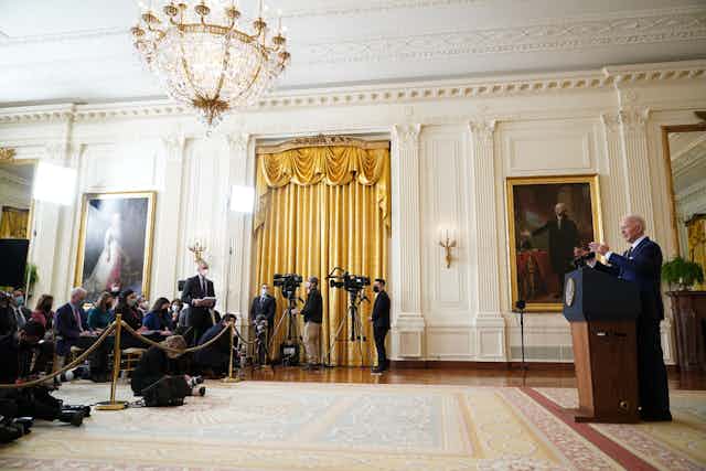 Joe Biden is seen from a distance, standing at a lectern and facing a row of people with cameras and notepads behind a rope.  