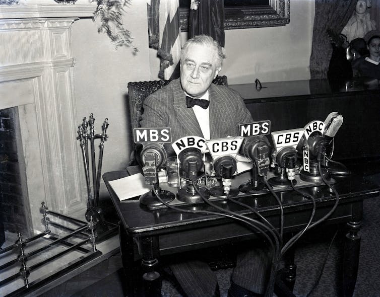 Franklin Delano Roosevelt sits at a table with microphones labeled 'CBS' and 'NBC' in a black and white photo.
