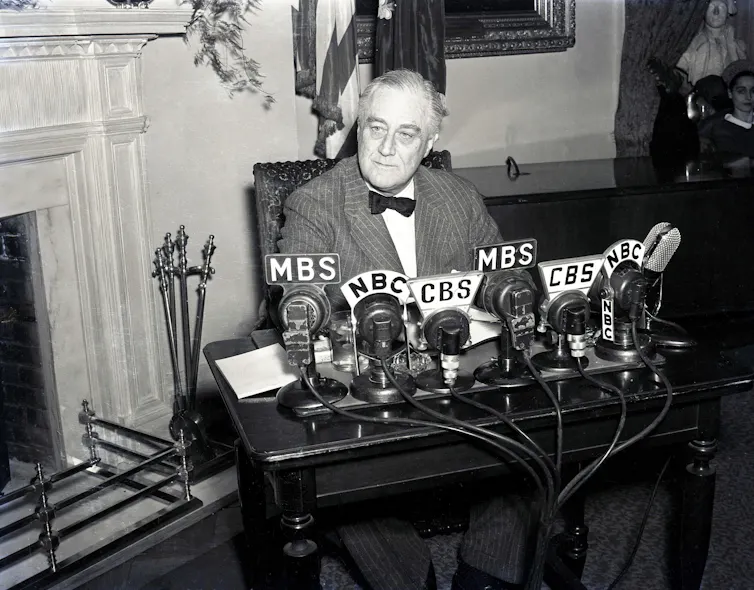 Franklin Delano Roosevelt sits at a table with microphones labeled 'CBS' and 'NBC' in a black and white photo.