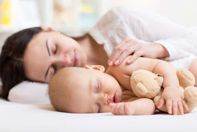A baby asleep on a bed, holding a stuffed toy, a woman asleep behind.