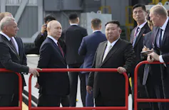 A balding man stands next to a rotund man behind a red metallic barrier. Both wear dark suits.