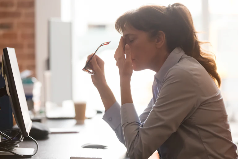 A woman sitting at a computer desk rubs her forehead with her eyes closed