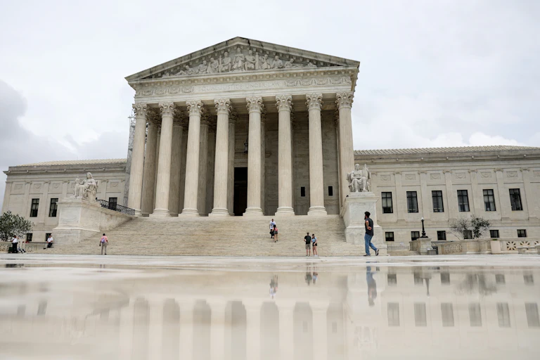 People walk on the landing and stairs of a large white building with pillars.