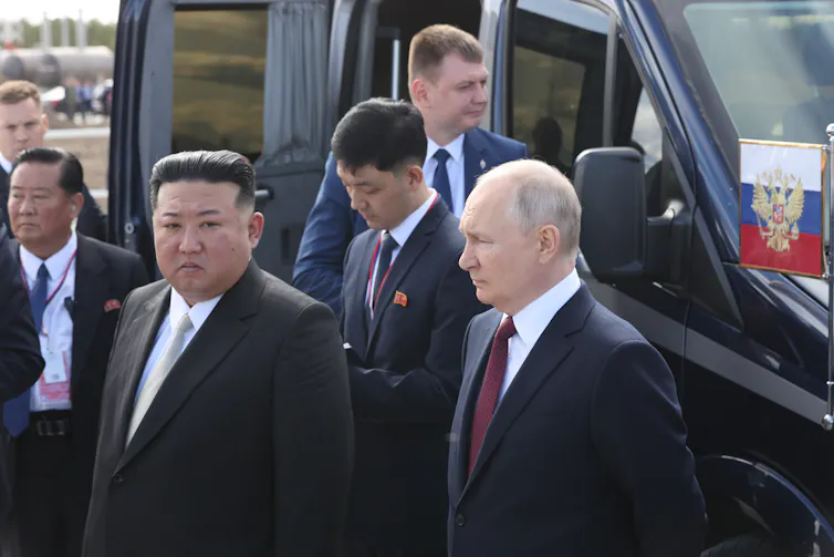 Two men. in suits stand next to a vehicle.