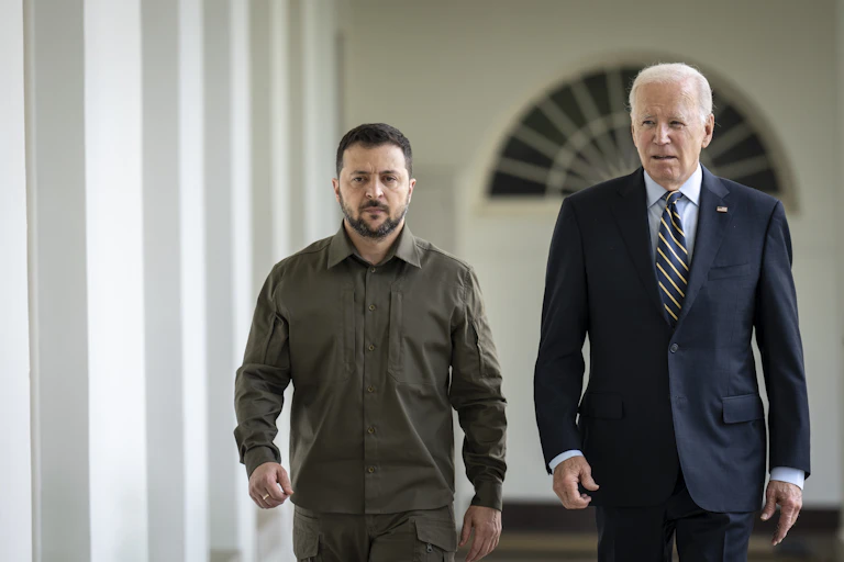A man in suit and tie walks with a bearded man in a grey/green shirt.