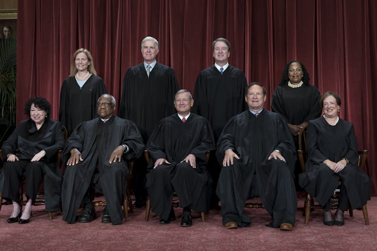 Nine people in black robes, sitting in two rows against a red curtain.