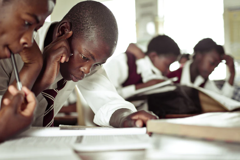 Two teenagers wearing white shirts with maroon and white striped ties pore over a book. There are other students doing the same in the background.