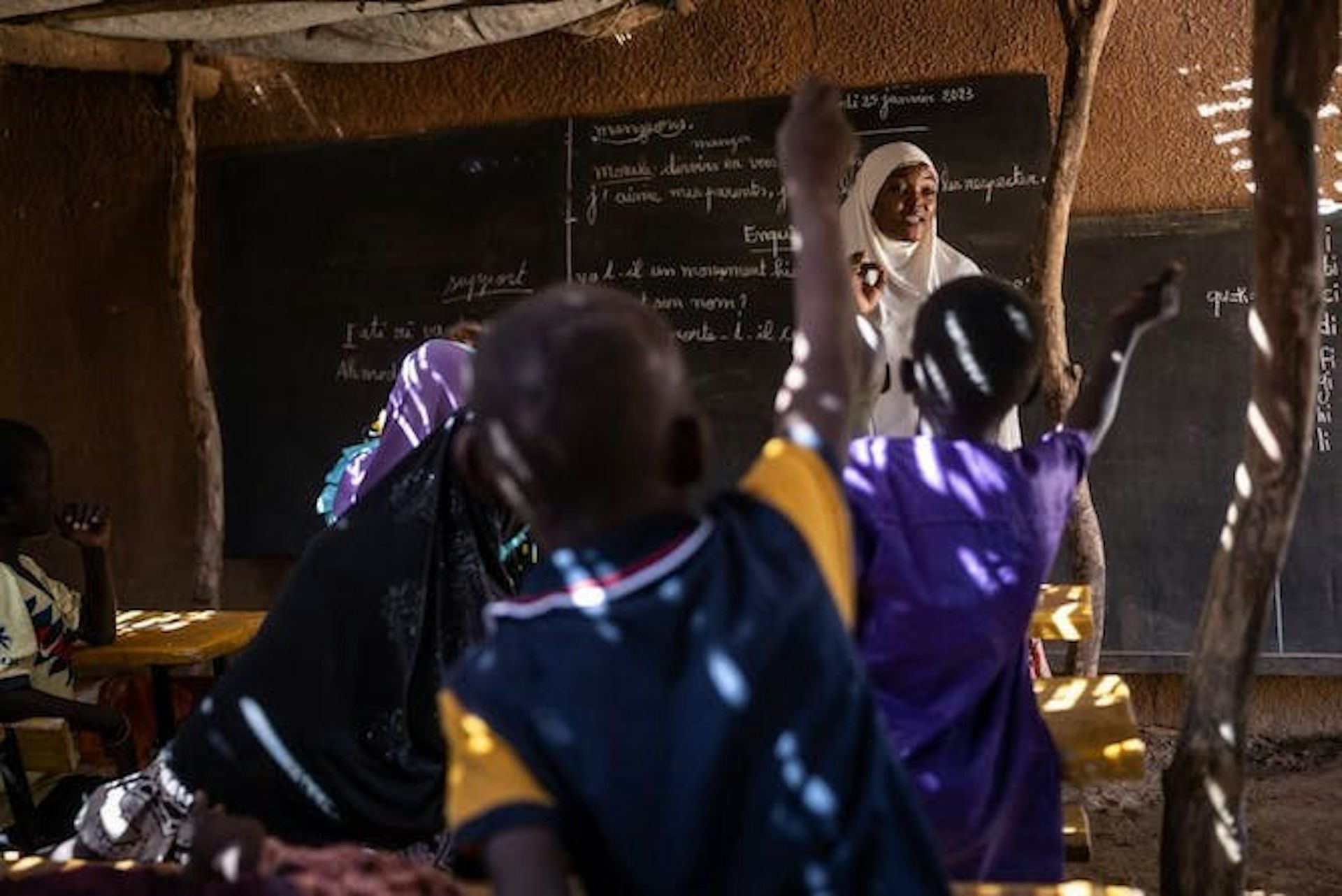 Une femme portant un hijab blanc se tient devant un tableau noir tandis que ses élèves lèvent la main