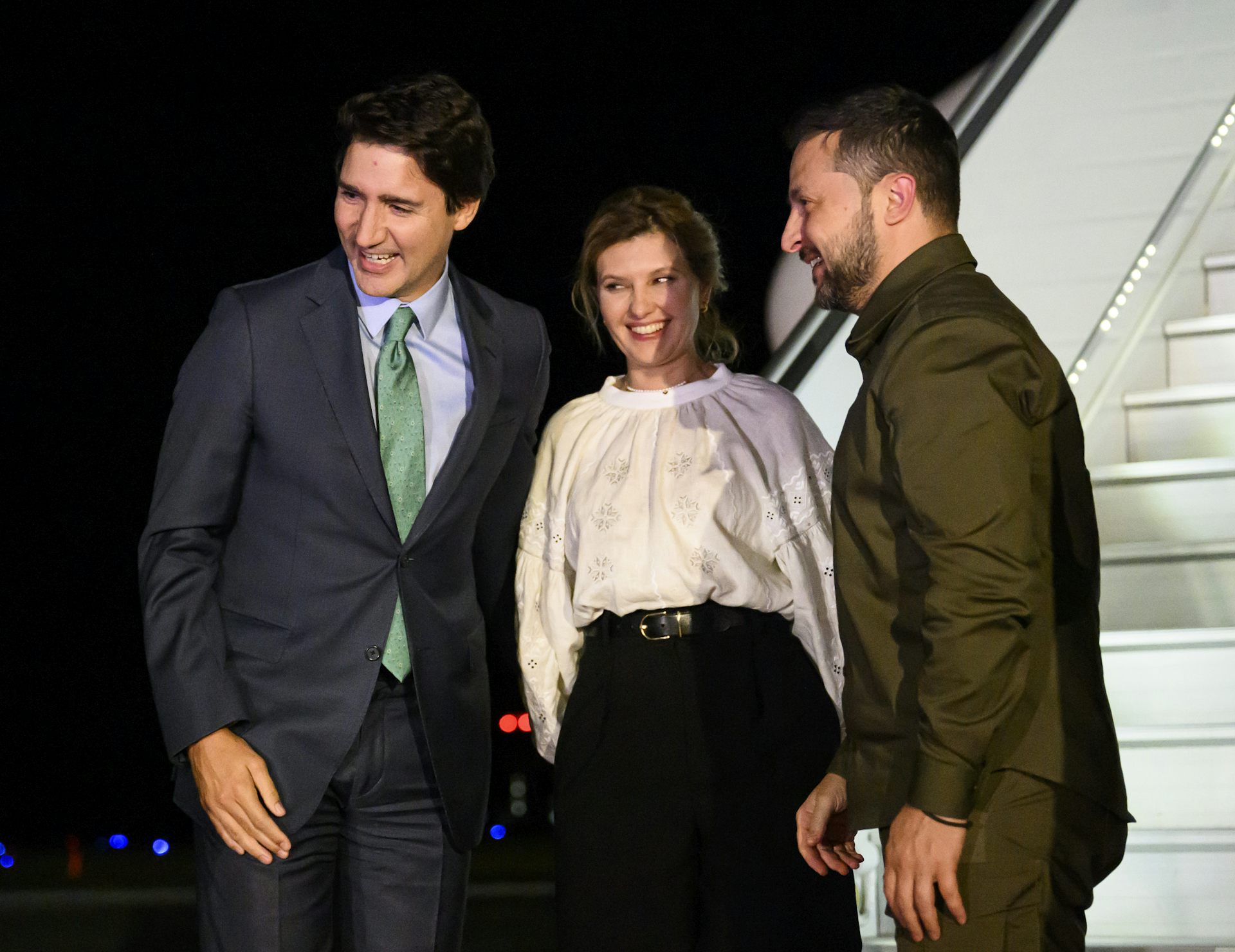 Two dark-haired men with a smiling woman between them smile as they stand on an airport tarmac beside a plane.