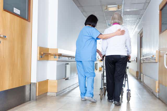 A woman with gray hair using a walker, assisted by a woman in blue scrubs, seen from behind in a long corridor