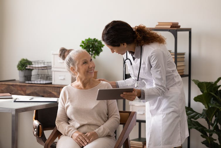 A woman in a white coat with a stethoscope and clipboard stands and talks to an older woman in a chair