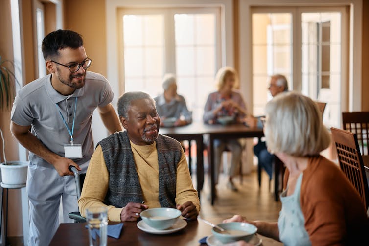 Two older women sitting at a table while a young man wearing an ID badge talks to them
