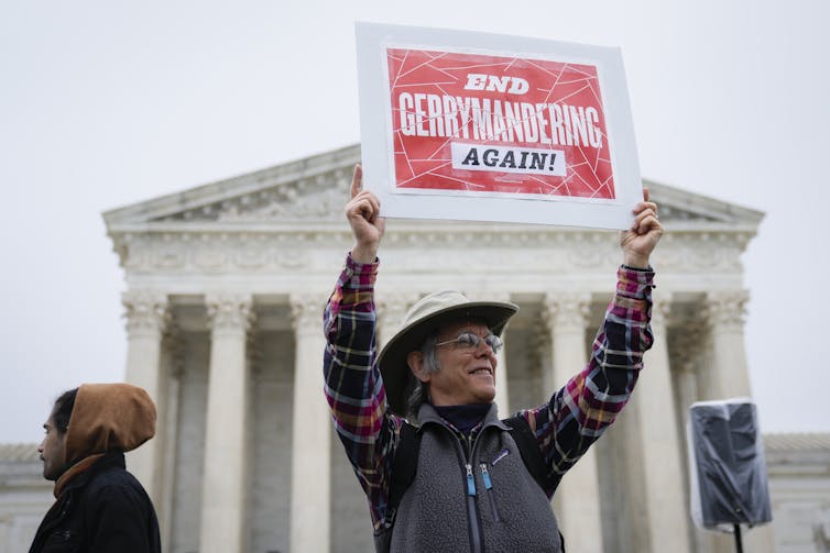 A man wearing a wide-brim, white hat, a red, black and white lumber jack shirt and grey vest stands before the US Supreme Court holding a sign that reads,