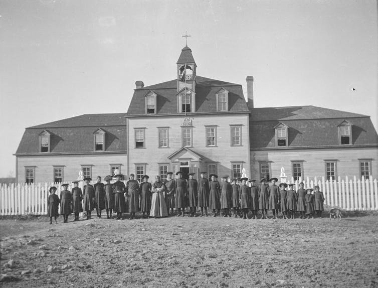 Children seen standing in rows in front of a 19th century institution with an adult.