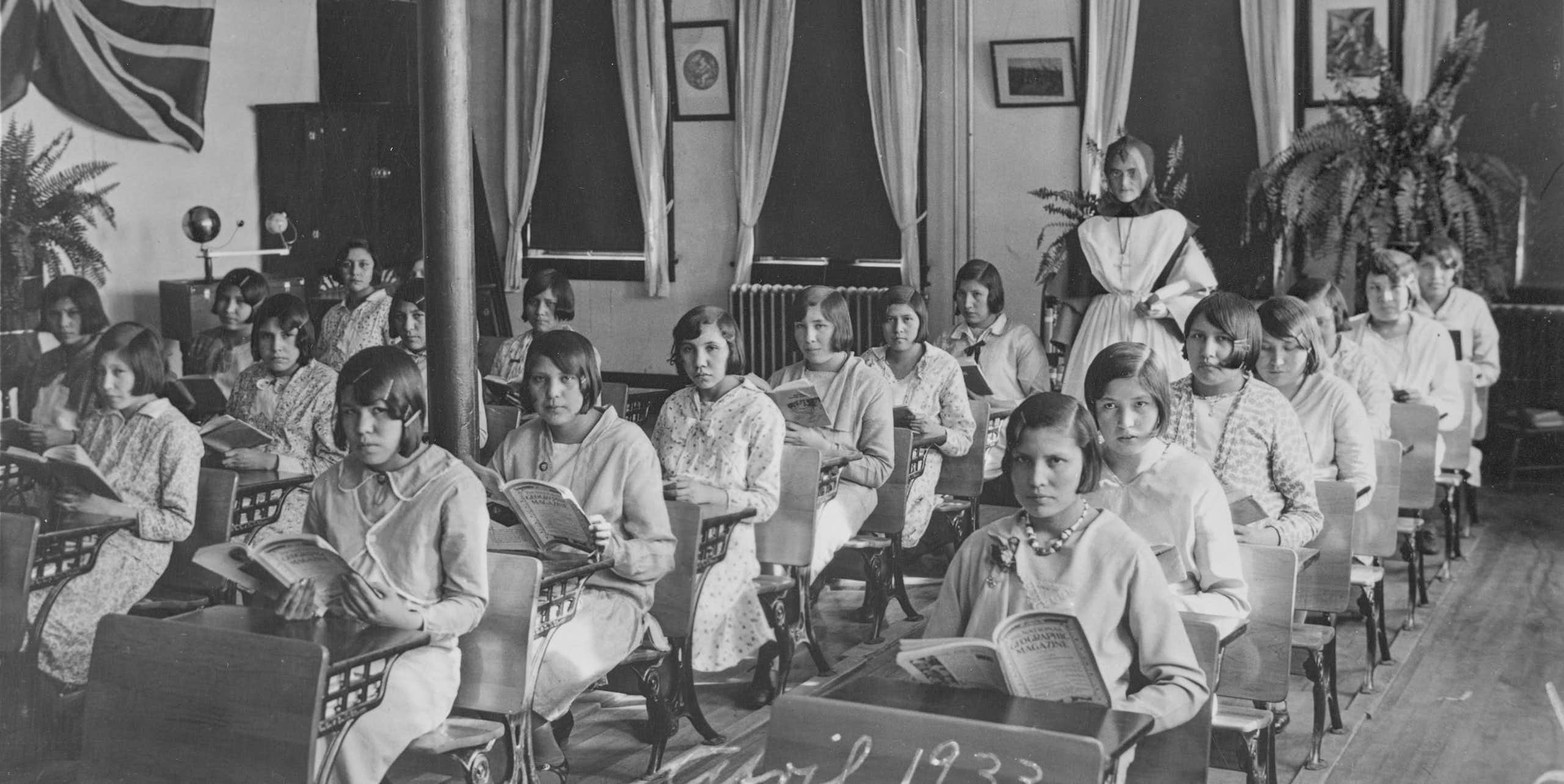 Black and white photo showing 1930s classroom of Indigenous adolescent girls sitting in rows of desks.