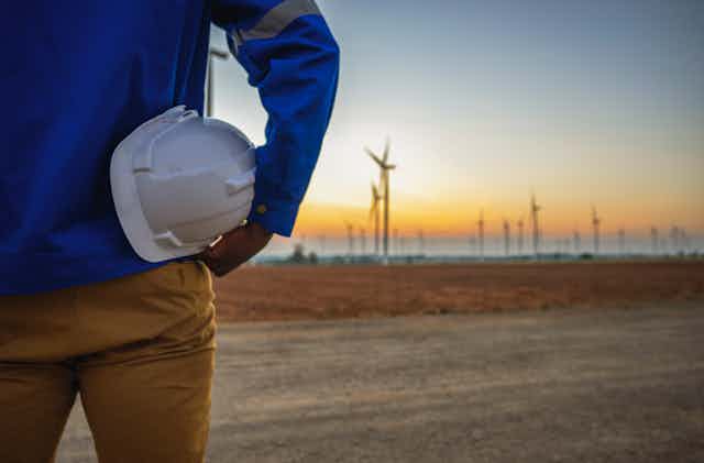 A person with hard hat under their arm stands looking at a wind farm in the distance.