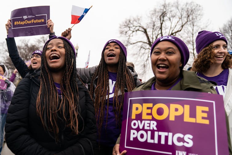 Five smiling women, each wearing purple winter hats, appear to also shout something in unison. One woman holds a sign that reads, 