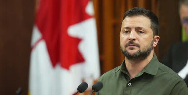 A bearded man in a dark green shirt stands in front of a podium with microphones. A Canadian flag is behind him.