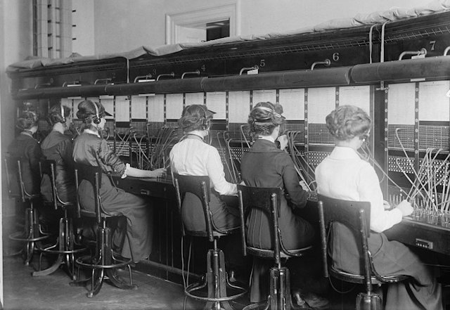 Black-and-white photo of six women wearing headphones and wearing old-fashioned clothing plugging cables into sockets